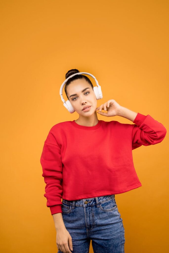 Portrait of a stylish young woman wearing white headphones and a red crop top against an orange background.