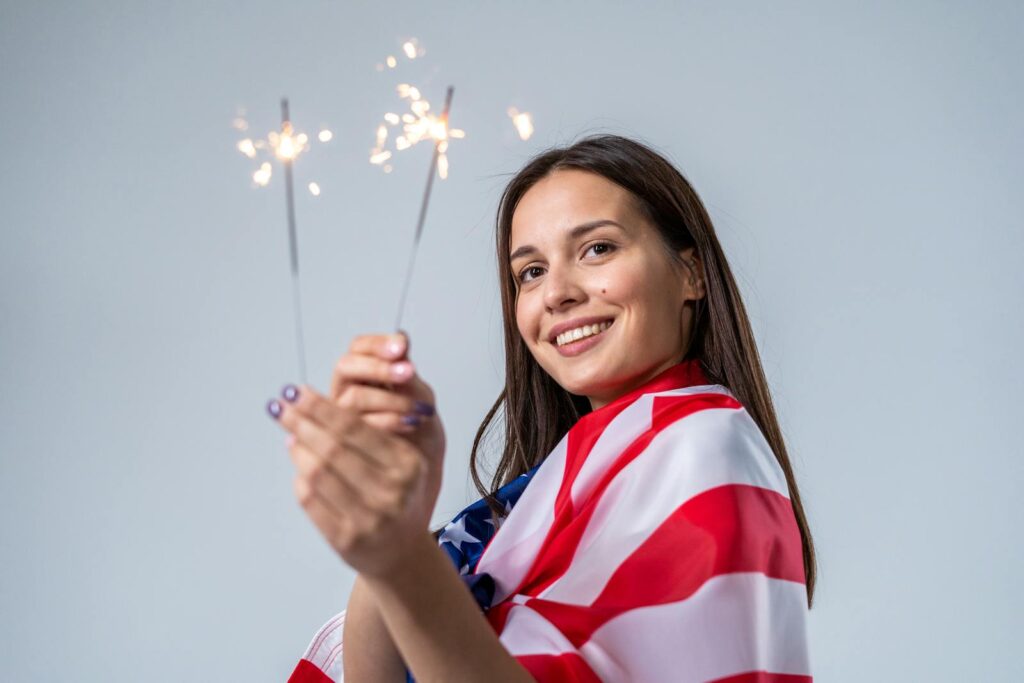 Young woman celebrating Independence Day with sparklers, wrapped in American flag.