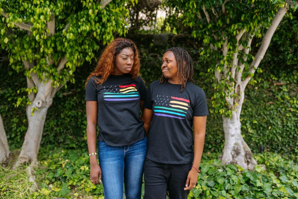 Two women smiling and embracing in matching pride t-shirts, standing outdoors among lush greenery.