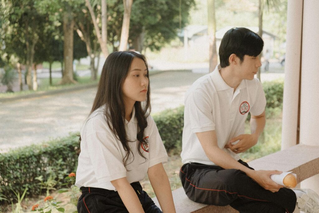 Two young adults sitting on a park bench in casual attire outdoors.