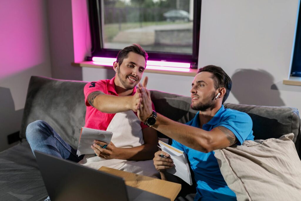 Two young men high-fiving while working on laptops at home, creating a collaborative study environment.