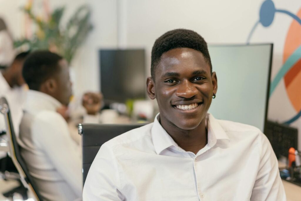 A cheerful young man in a white shirt smiling at his desk in a modern office environment.