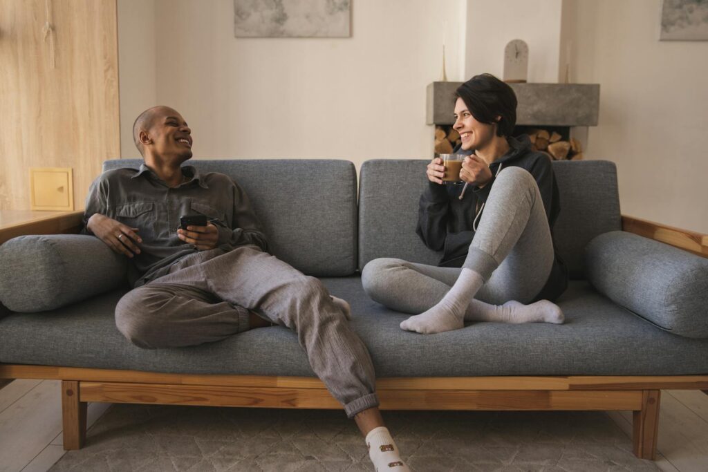 Positive young multiracial couple resting on sofa and talking to each other while enjoying weekend in cozy living room at daytime