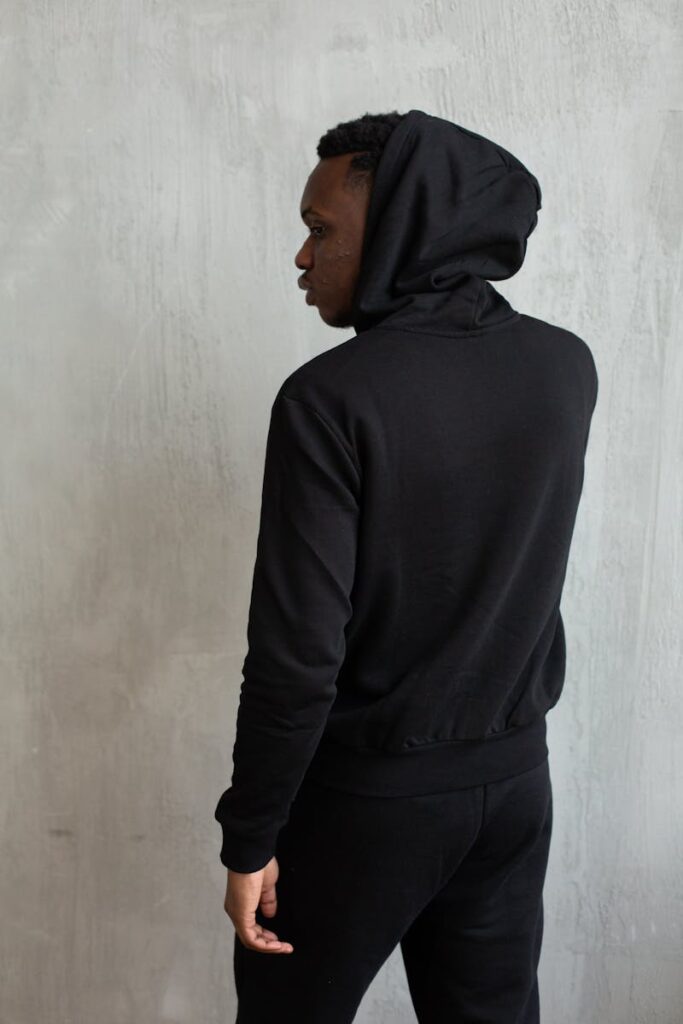 Back view of African American male in sports suit with hood on head standing against gray background