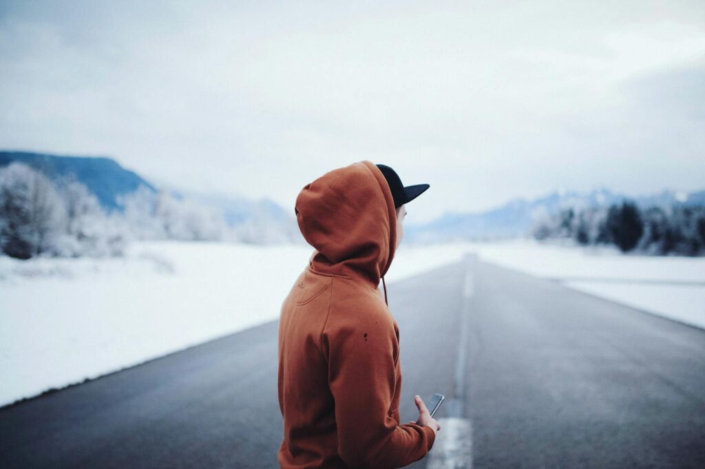 A person in a hoody stands on a snowy road, holding a smartphone, with mountains in the background.