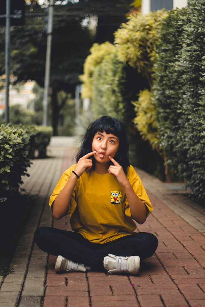 Young woman in a yellow shirt poses playfully on a city sidewalk with greenery, expressing joy.