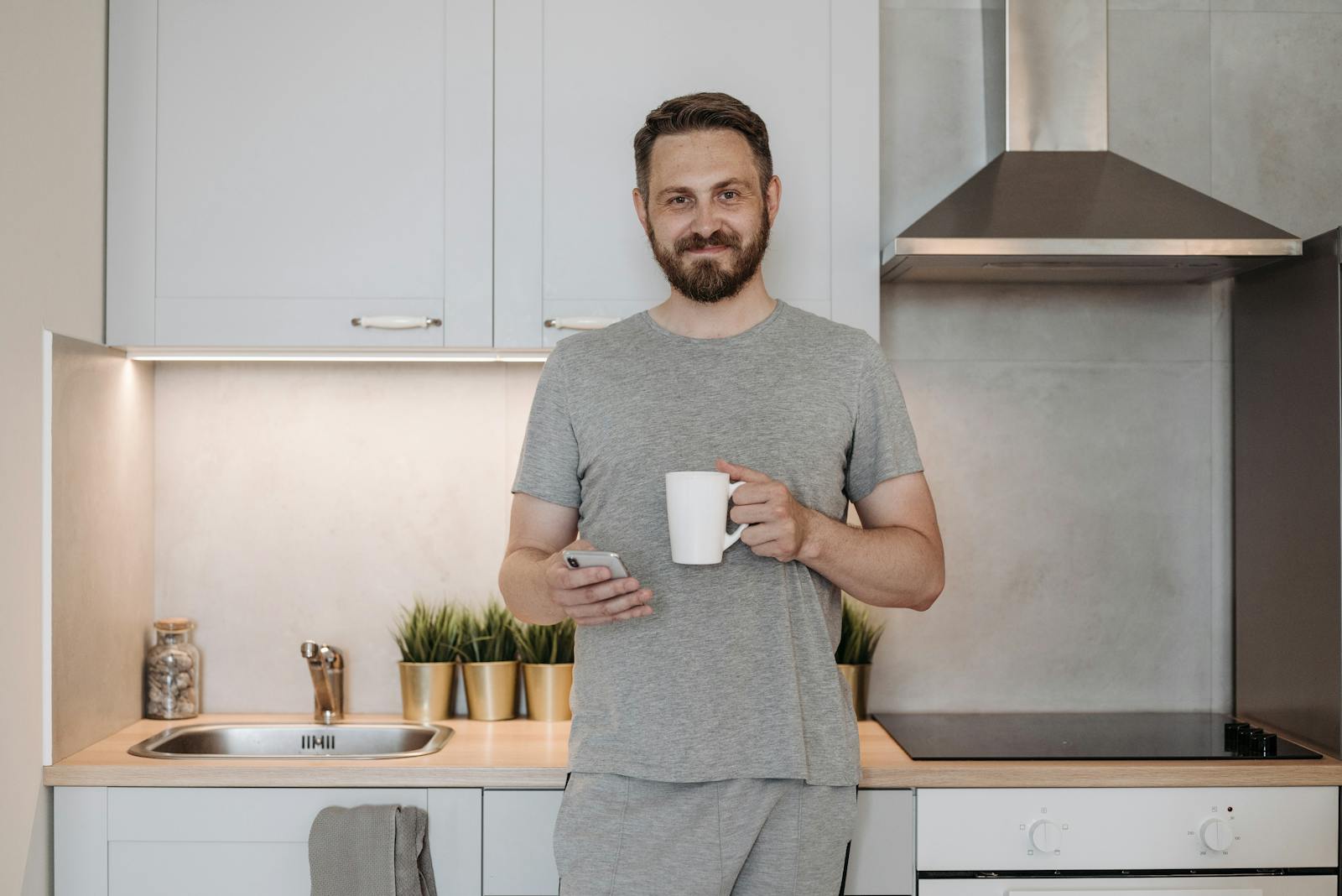 A bearded man in a gray shirt holding a mug and smartphone in a modern indoor kitchen. comfort colors t shirts