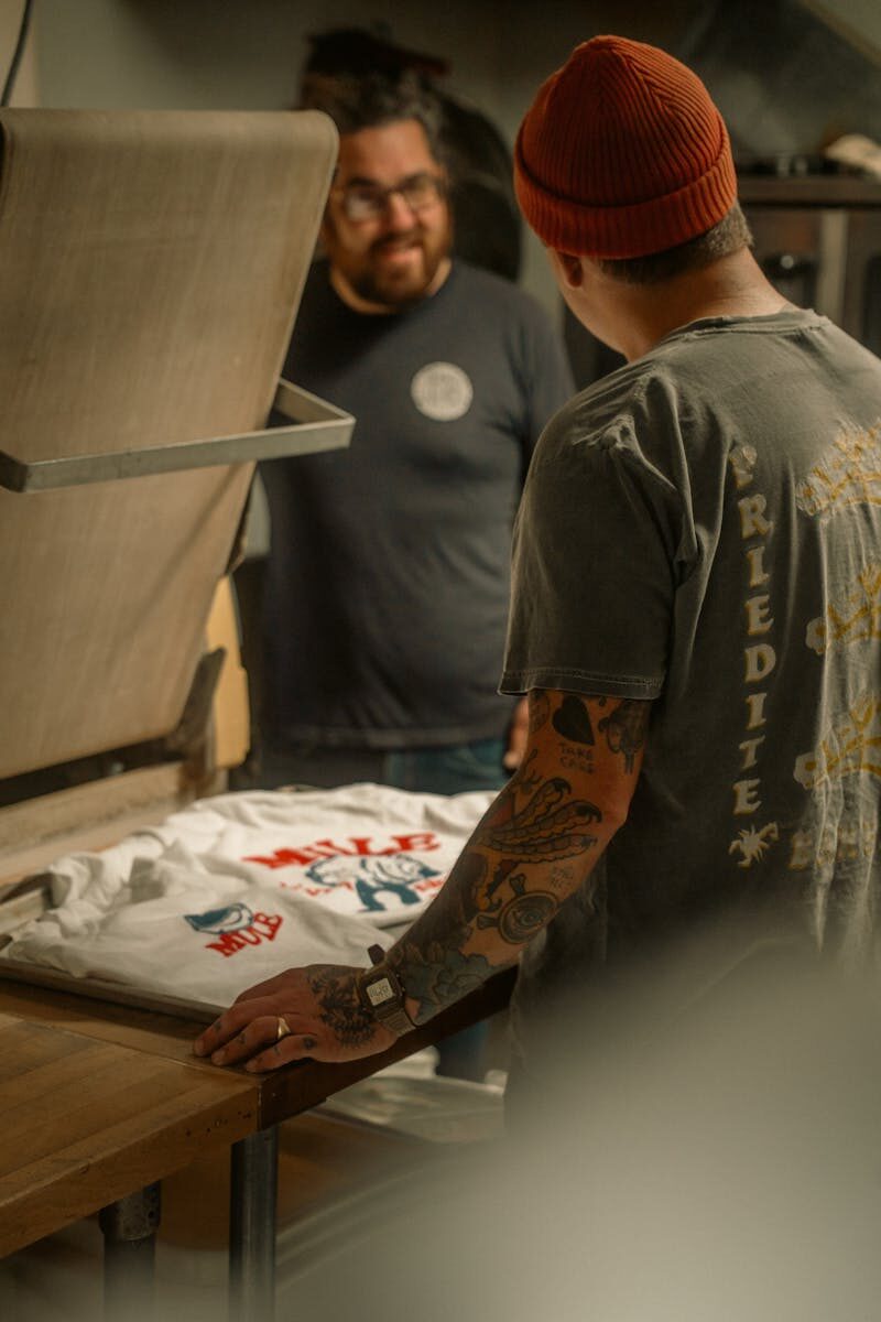 Photo by James Collington Two men engaged in screen printing a t-shirt in a cozy workshop setting.