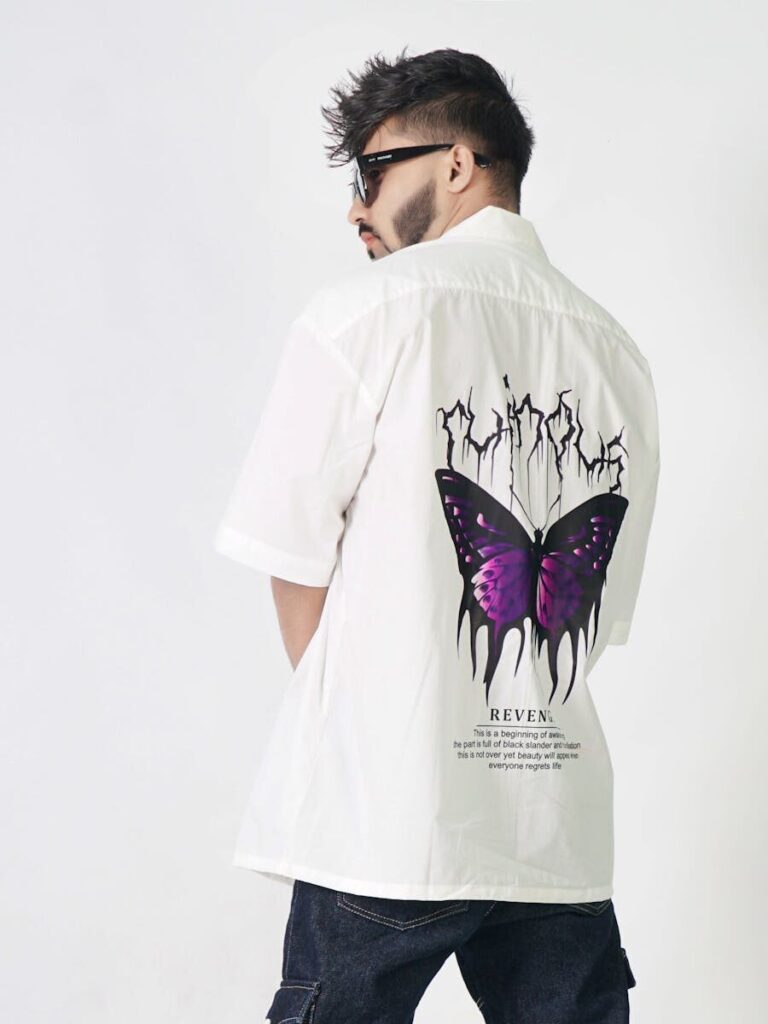 Stylish man wearing sunglasses and a shirt with purple butterfly design standing against a white background.