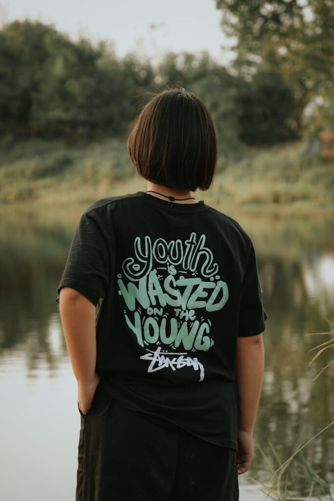 Back view of person standing by a peaceful lake surrounded by trees in summer, wearing a graphic tee.