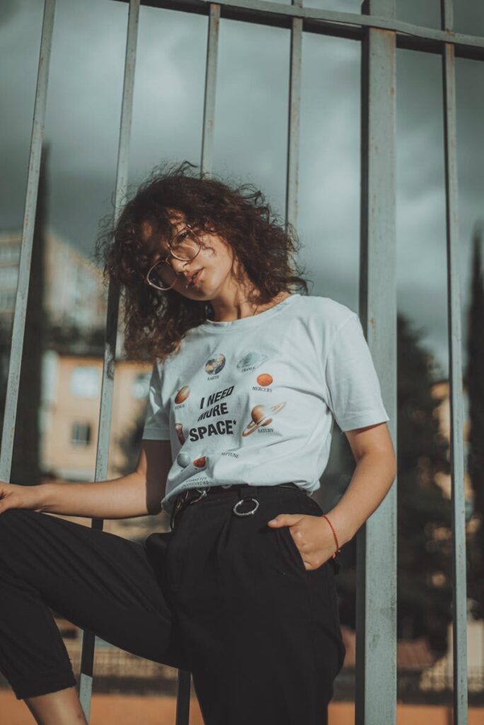 Hip young woman with curly hair poses against a fence in an urban Russian landscape.