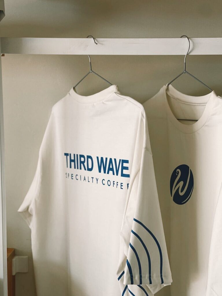 White t-shirts hanging with 'Third Wave Specialty Coffee' branding. Minimalist design.