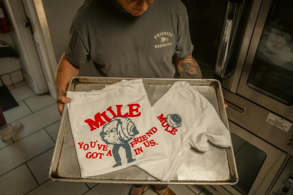 Person showcasing white T-shirts with red mule design and humorous text, indoors. Direct to Film Transfers Explained: How They Work and Why They’re So Popular