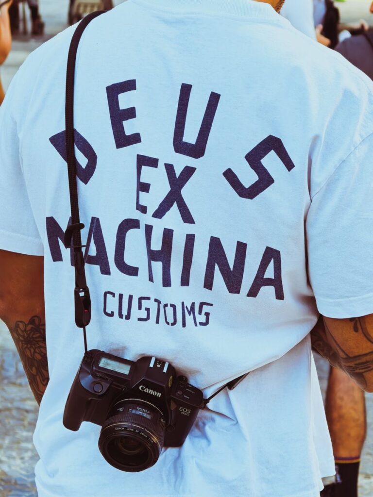 Back view of a man wearing a 'Deus Ex Machina' t-shirt with camera gear.